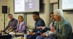 Five individuals are seated on chairs in front of an audience during a panel discussion. Each panelist holds a microphone.